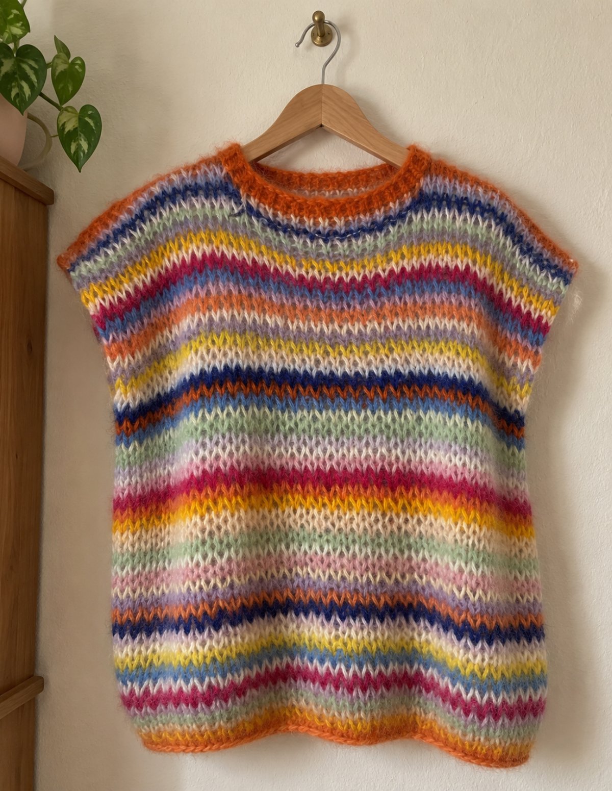 Colourful striped mohair slipover vest with an orange neckband, hanging on a wooden hanger against a white wall