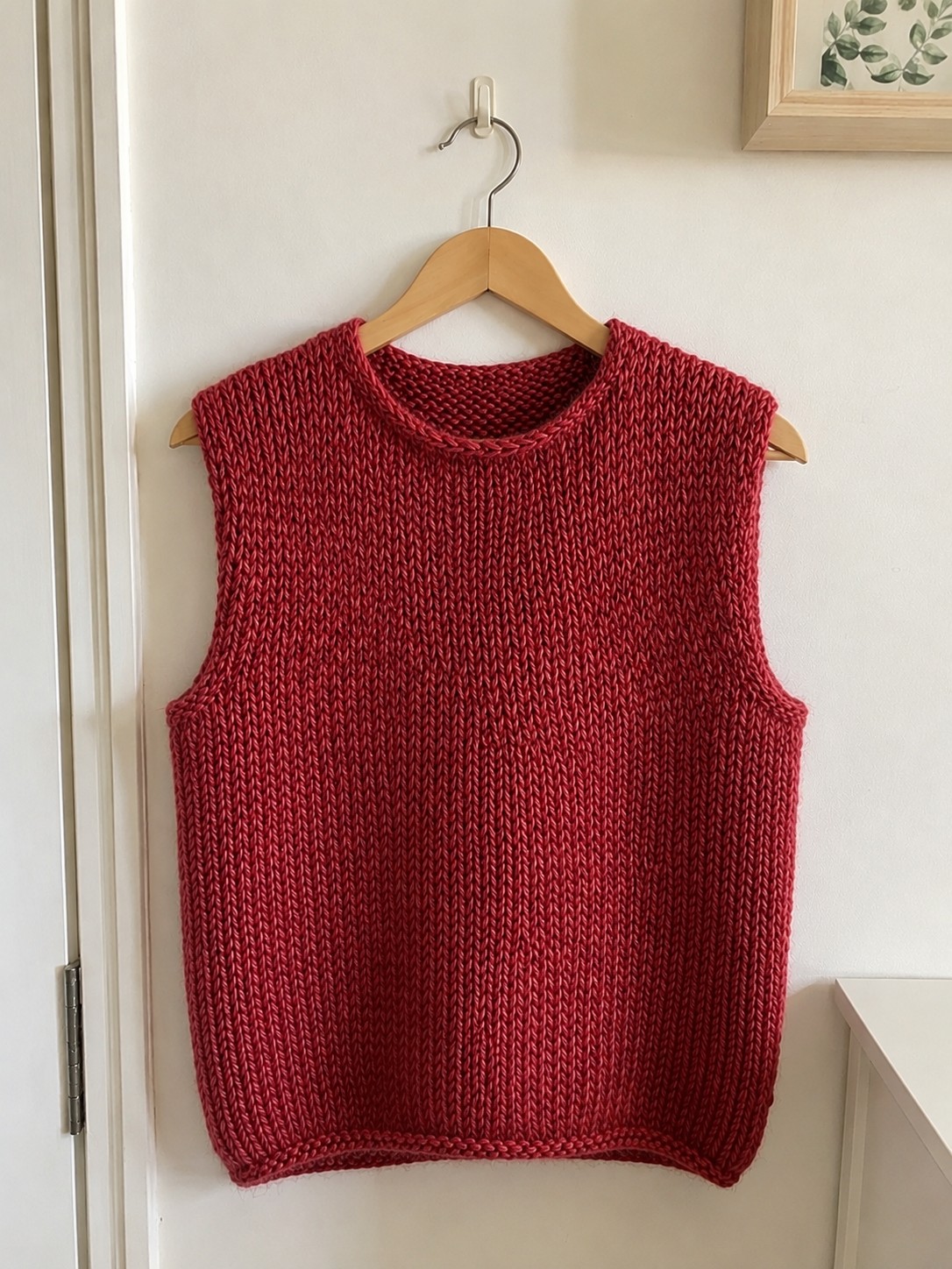 Longline knitted vest in deep rosewood red with rolled stockinette edges, hanging on a wooden hanger